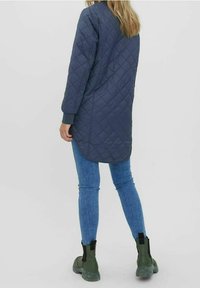 Kvadderad marinblå lång kappa med räfflade manschetter, i kombination med ljusblå figurnära jeans och gröna ankellånga stövlar. Enkel design utan synliga mönster.