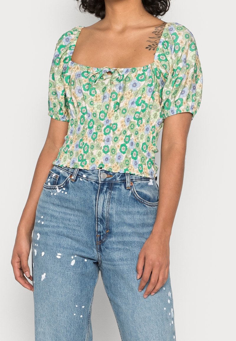 Femme portant un chemisier vert à manches bouffantes fleuries et un jean bleu taille haute avec des éclaboussures de peinture blanche.