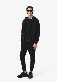 Tenue de sport noire comprenant un sweat à capuche zippé et un jogging ajusté. Tissu texturé, couleur unie, assortie à des baskets noires et des lunettes de soleil.