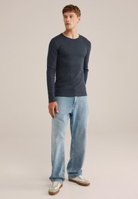 Jaunas vyras stovi, dėvėdamas tvirtą tamsią ilgomis rankovėmis marškinį, šviesiai mėlynas laisvas džinsus ir baltus sportbačius, žiūrėdamas į šoną.