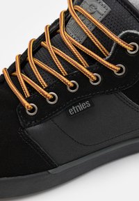 Sapatos de skate em camurça preta e sintética apresentam atacadores trançados amarelos e roxos, ilhoses de metal e um logótipo costurado na lateral.