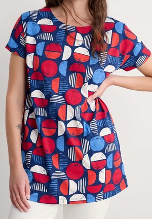 Woman wearing short-sleeve navy blue shirt with red, white, and light blue geometric circles and half-circles, hand on hip.