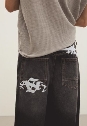 Persoană care poartă pantaloni negri din denim cu imprimeu mare alb în stil graffiti pe buzunarele din spate și un top bej fără mâneci, tricotat cu model tip vafe.