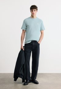 Νέος άνδρας που φοράει ανοιχτό μπλε κοντομάνικο πουκάμισο, μαύρο τζιν, μαύρα παπούτσια, κρατώντας ένα μαύρο σακάκι στο ένα χέρι, στέκεται σε γκρι δάπεδο.