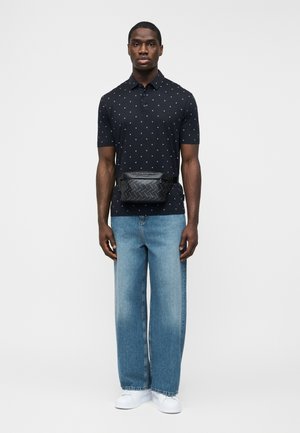 Férfi fekete mintás galléros pólóban, világoskék bőszárú farmernadrágban, fehér sportcipőben és fekete, geometriai mintás deréktáskával.