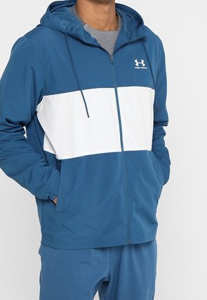 Personne portant une veste à capuche zippée bleu et blanc, les mains dans les poches, associée à un pantalon bleu assorti.
