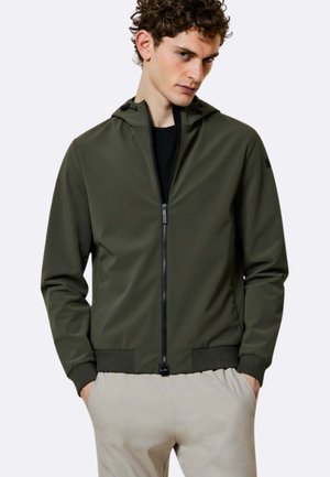 Uomo che indossa una giacca con cappuccio verde scuro con zip e polsini a costine, e pantaloni beige, in piedi con le mani nelle tasche davanti a uno sfondo neutro.