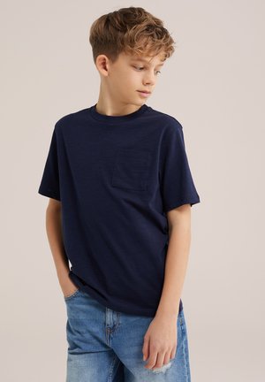 Niño con cabello rizado que lleva una camiseta azul marino y vaqueros rasgados azul claro, manos en los bolsillos, mirando hacia su izquierda sobre un fondo liso.