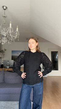 Fekete blúz, finom hímzett részletekkel, puffadt ujjak és gombos záródás jellemzi, széles lábú kék farmernadrággal párosítva.
