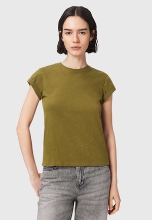 Jeune femme aux cheveux courts et foncés, portant une chemise courte à manches courtes couleur vert olive et un jean gris, debout devant un fond uni.