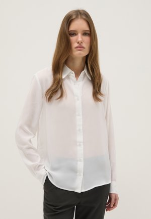 Joven mujer con cabello largo y castaño, vistiendo una blusa blanca transparente de botones y pantalones negros, de pie contra un fondo claro y sencillo.