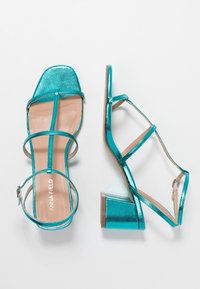 Turkoosi korkokengät metallipinnalla, avonainen neliskulmainen varvas ja useita kapeita hihnoja. Tukevan muotoilu varsi vakauden takaamiseksi.