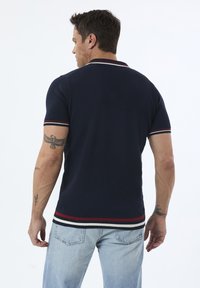 Marineblaues Poloshirt mit strukturiertem Muster, kontrastierenden roten und weißen Streifen am Kragen und an den Ärmeln, kombiniert mit hellblauen Jeans.
