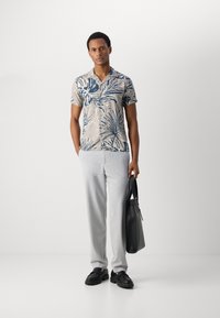Cămașă cu mânecă scurtă în bej, cu imprimeu tropical albastru, asortată cu pantaloni gri deschis și pantofi negri, purtând o geantă tot neagră.