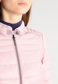 Vaaleanpunainen tikattu takki, jossa on pystykaulus, koko vetoketju ja napin kiinnitys; ominaisuuksiin kuuluu vaakasaumojen ompelu ja sileä rakenne.