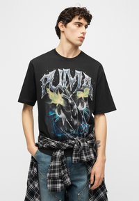 Νέος άνδρας με σγουρά μαλλιά, φοράει μαύρο γραφικό t-shirt Puma και τζιν, με ένα πουκάμισο καρό δεμένο γύρω από τη μέση του, κοιτάζοντας πλάγια.