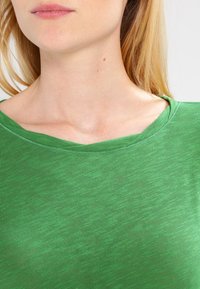 T-shirt verde, leggero, con una texture morbida, caratterizzato da un ampio scollo e maniche corte. Il tessuto presenta una delicata fantasia melange.