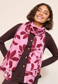 Bufanda rosa con patrones de hojas burdeos oscuros, hecha de una tela ligera, llevada sobre una blusa oscura con botones dorados.
