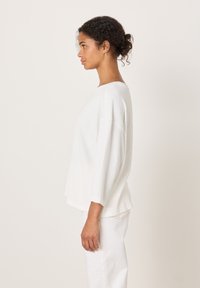 Kvinne med krøllete hår i en knute, iført en løs hvit langermet topp og hvite bukser, stående i profil mot en nøytral bakgrunn.