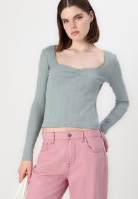 Vaaleanvihreä pitkähihaisessa paidassa on neliömäinen kaula-aukko, tekstuuriuritus ja keskeinen poimu. Yhdistetty vaaleanpunaisiin ja valkoisiin raidoitettuihin farkkuihin.