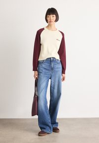 Woman stands wearing cream and burgundy long-sleeve sweater, wide-leg blue jeans, burgundy shoes, and holding a burgundy handbag by her side.