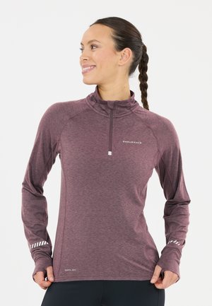 Vrouw met een gevlochten paardenstaart die een bordeauxrode sporttop met lange mouwen en duimgaten draagt, staat zijwaarts en glimlacht.