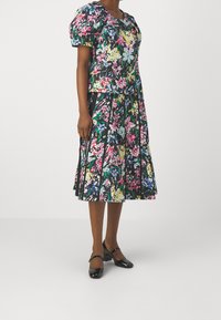 Abito stampato floreale con corpino aderente, maniche a sbuffo e gonna plissettata. Sfondo nero con motivi floreali colorati. Scarpe nere con tacco.