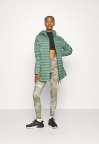 Hellgrüne gepolsterte Jacke mit Kapuze, schwarzes Cropped-Top, beige Camo-Leggings und schwarze Sportschuhe. Glatte Textur und passgenaues Design.