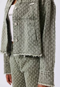 Groene denimjas met een gestructureerd bloemmotief, rafelige randjes en zilveren drukknoopjes, gecombineerd met bijpassende denim broek met textuur.