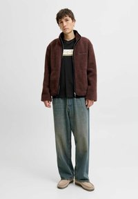 Vinsdæmpet fleecejakke med oversized pasform, frontlommer og lynlås, båret over en sort grafisk t-shirt og slidte brede jeans.