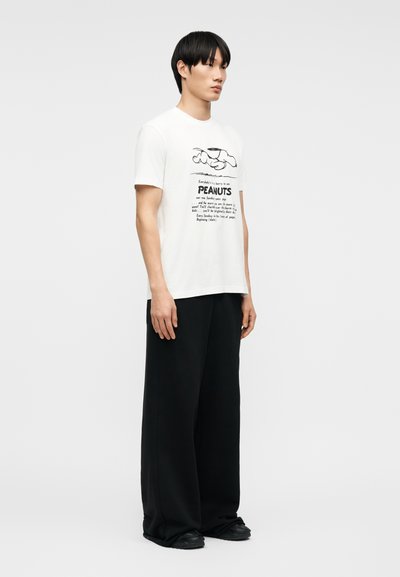 Νεαρός άνδρας που στέκεται φορώντας λευκό μπλουζάκι με σχέδιο Peanuts, μαύρο παντελόνι με φαρδύ πόδι και μαύρα παπούτσια σε απλό ανοιχτό φόντο.
