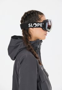 Hiihtolasit mustalla hihnalla, jossa on "SLOPE"-logo, peililinssit ja tiukka istuvuus. Henkilöllä on päällään harmaa hupparitakki.