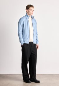 Ανοιχτό μπλε μπουφάν με φερμουάρ, φτιαγμένο από απαλό ύφασμα, φορεμένο πάνω από ριμπ μπεζ πουλόβερ. Συνδυασμένο με μαύρο παντελόνι και μαύρα παπούτσια.