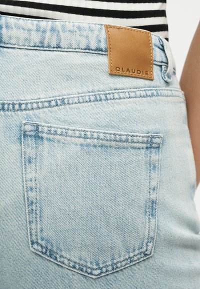 Φωτεινά μπλε τζιν παντελόνια με πίσω τσέπη και καφέ δερμάτινο έμβλημα με την επιγραφή "CLAUDIE", φορεμένα με μαύρο-άσπρο ριγέ τοπ.