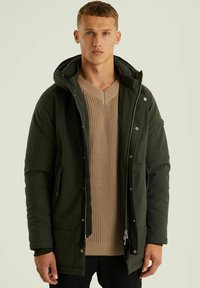 Veste étanche verte avec capuche, fermée par zip et boutons-pression, associée à un pull en maille beige texturé en dessous.