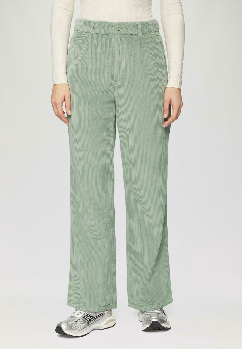 Femme portant un pantalon en velours côtelé vert clair, un haut blanc à manches longues côtelées et des chaussures de sport argentées, debout devant un fond uni.