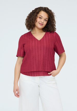 Kobieta o kręconych włosach, ubrana w czerwoną, teksturowaną bluzkę z krótkim rękawem i dekoltem w serek oraz białe spodnie z szerokimi nogawkami, stojąca z jedną dłonią w kieszeni.