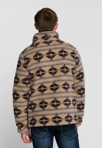 Beige fleecetrui met geometrische patronen in bruin, blauw en crème; voorzien van een hoge kraag en lange mouwen, wat een gezellige textuur biedt.