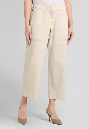 Donna che indossa pantaloni beige cropped a gamba larga e tacchi slingback taupe con punta appuntita, in piedi davanti a uno sfondo grigio.