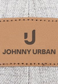 Lähikuva ruskeasta nahkalapusta, jonka reunat on ommeltu harmaalle kankaalle, jossa on tyylitelty J-kirjain ja teksti "JOHNNY URBAN."