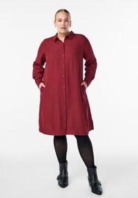 Červené pruhované šaty košilového strihu s gombíkmi vpredu, dlhými rukávmi, bočnými vreckami a voľným strihom. Čierne členkové čižmy s dekoračnými prvkami.