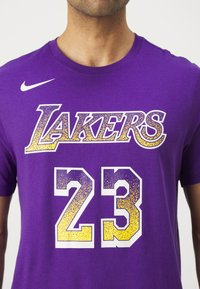Violetinio medvilninio marškinėlių su atspausdintu "LAKERS" baltos ir geltonos stilizuotos spalvų kombinacijoje, o žemiau esantis numeris "23" turi gradientinį dizainą. Nike logotipas ant rankovės.