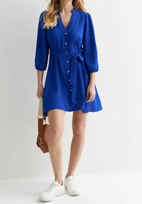 Abito blu con scollatura a V, chiusura con bottoni e vita aderente. Presenta maniche a sbuffo e una vestibilità comoda, abbinato a sneakers bianche.