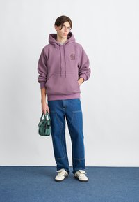Lilla fleece hettegenser med brodert logo på brystet, kombinert med blå jeans og en liten grønn håndveske. Hvit sneakers fullfører antrekket.