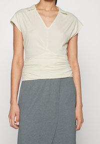 Cream short-sleeve top with a v-neck and collar; features a ruched detail at the waist, paired with a gray skirt. Lightweight fabric.