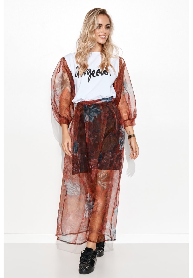 Biała koszulka z napisem "gorgeous." w towarzystwie zwiewnej, półprzezroczystej spódnicy maxi w kolorze rdzy z kwiatowym wzorem. Czarne sneakersy z ozdobami.