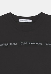 Czarna bawełniana koszulka z okrągłym dekoltem i poziomym logo Calvin Klein Jeans w kontrastującym białym kolorze na pasku na wysokości klatki piersiowej.