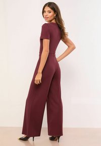 Bordó overál rövid ujjakkal, testhezálló felsőrésszel és bő szárú nadrággal. Simakötött anyagból készült, hátul cipzáras részlettel és minimális fém kiegészítőkkel.