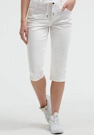 Person trägt weiße gekürzte Hosen mit Kordelzug in der Taille und weiße Sportschuhe, steht vor einem einfarbigen hellen Hintergrund.