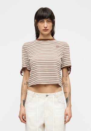 Femme portant un T-shirt court à rayures marron et blanches avec des manches nouées et un pantalon blanc taille haute, debout devant un fond uni.